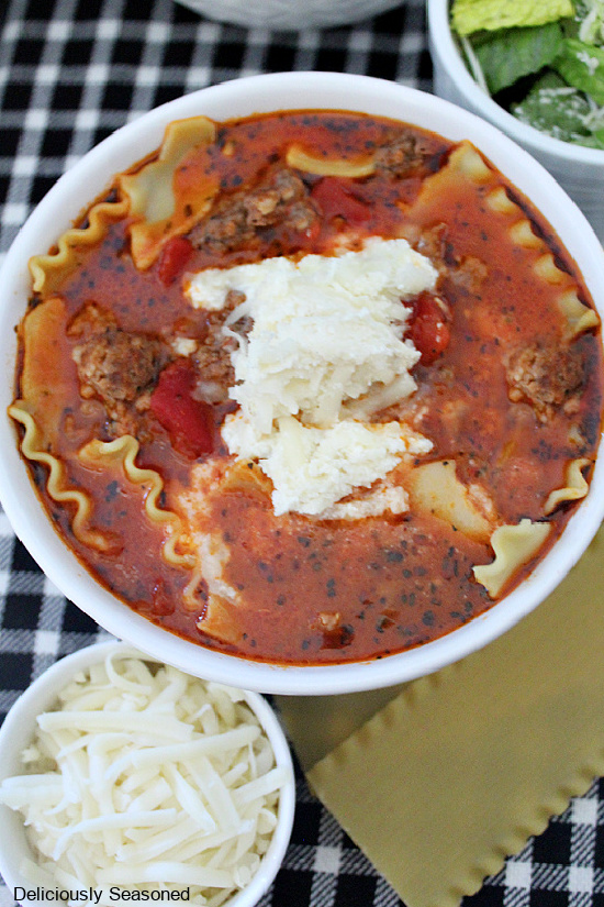 A white bowl filled with lasagna soup with a side salad, and a small bowl of mozzarella cheese placed on a black and white placemat.