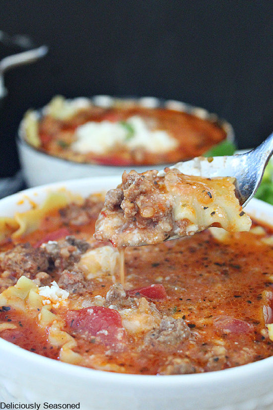A white bowl of lasagna soup with a spoonful of soup being held above the bowl of soup. Another white bowl filled with soup in the background.
