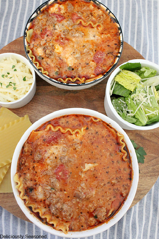 A round brown cutting board with two bowls white bowls of lasagna soup, two small white bowls, one with a side salad and one with shredded Mozzarella cheese, with uncooked lasagna noodles laying next to the bowl of soup.