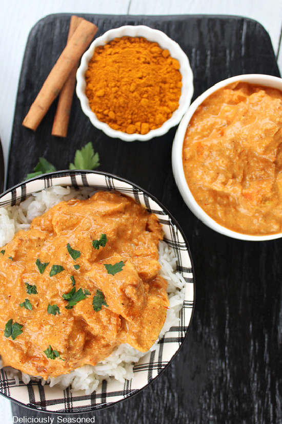 An overview picture of Butter Chicken in a black and white plaid bowl, a small white bowl filled with garam masala, and a white bowl filled with Butter Chicken with cinnamon sticks and cilantro in between bowls.