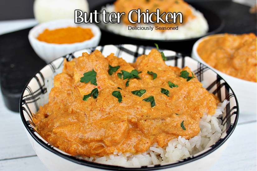 Butter Chicken over rice in a black and white plaid bowl with an onion, spices, and butter chicken  in the background.