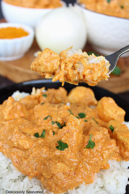 Butter chicken in a black bowl served over rice with a fork full of chicken over the bowl with an onion and two more bowls full of butter chicken in the background.