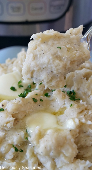 A close up picture of a heaping spoonful of mashed potatoes held up over a bowl of mashed potatoes.