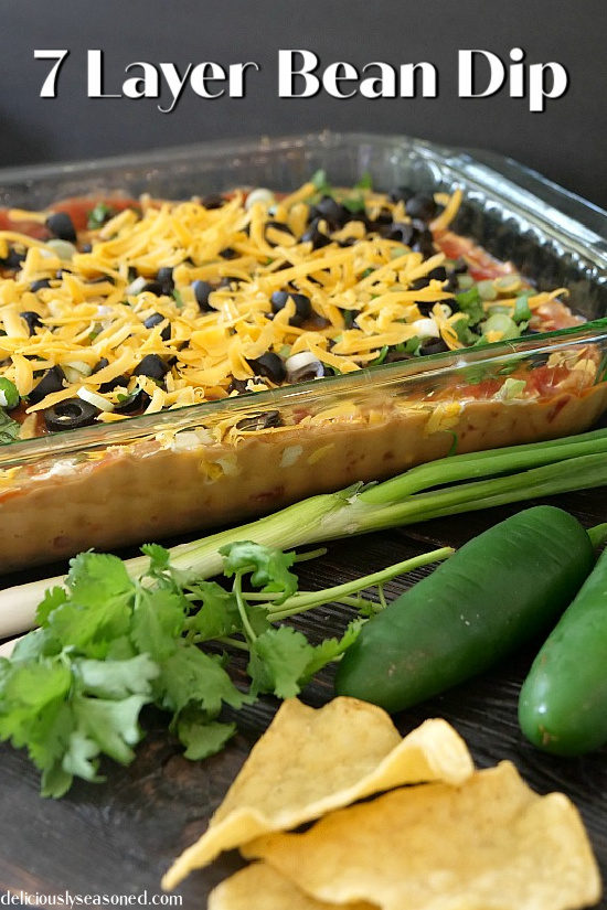 7 layer bean dip in a glass pan with cilantro, green onions, jalapenos, and chips in the foreground for decoration.