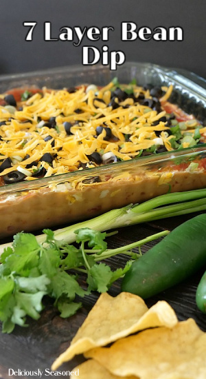 7 Layer bean dip in a glass pan with green onions, a jalapeno, cilantro, and chips in front of the pan.