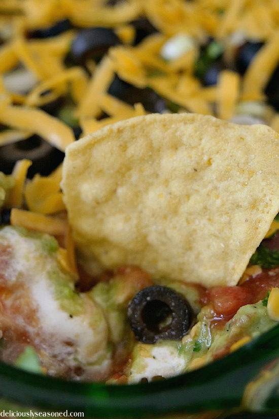 A close up photo of 7 layer dip with cheese, olives, sour cream, guacamole, refried beans, and salsa a chip dipped in it. 
