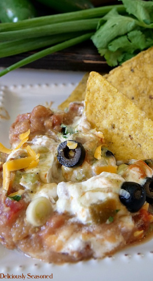 A serving of 7 layer bean dip on a white plate with chips on the side and green onion and cilantro in the background.