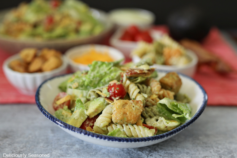 A white bowl with blue trim filled with lettuce, tomatoes, bacon, avocado, croutons, cheese and rotini pasta and another salad in the background.