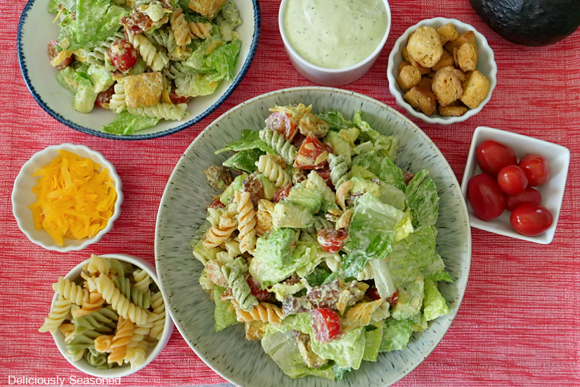 Two bowls with BLT Avocado Pasta Salad in it with all the ingredients in small bowls around the bigger bowls of salad.