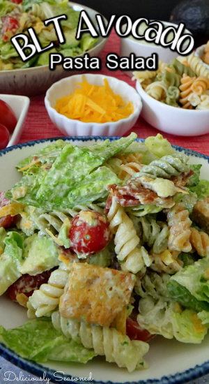 A white bowl with blue trim with BLT avocado pasta salad in it with grated cheese, pasta and another salad in the background.