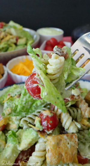A close up view of a fork with lettuce, pasta, tomato and bacon being held up over the bowl of salad showing the ingredients in the salad.