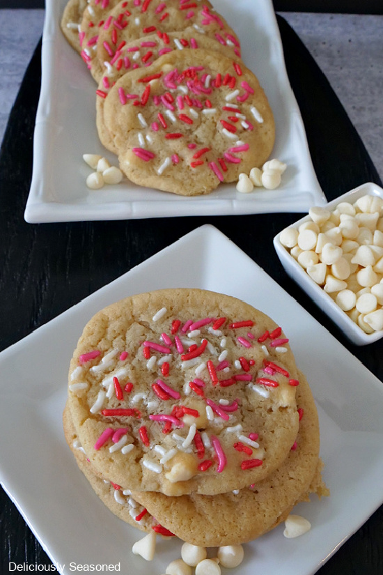 A black cutting board with 2 white plates on it with sugar cookies and white chocolate chips scattered on the plate, with a small white bowl of white chocolate chips.