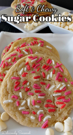 A white plate with 4 sugar cookies laying on it, with a small white bowl of white chocolate chips and a white plate with sugar cookies stacked on it in the background.