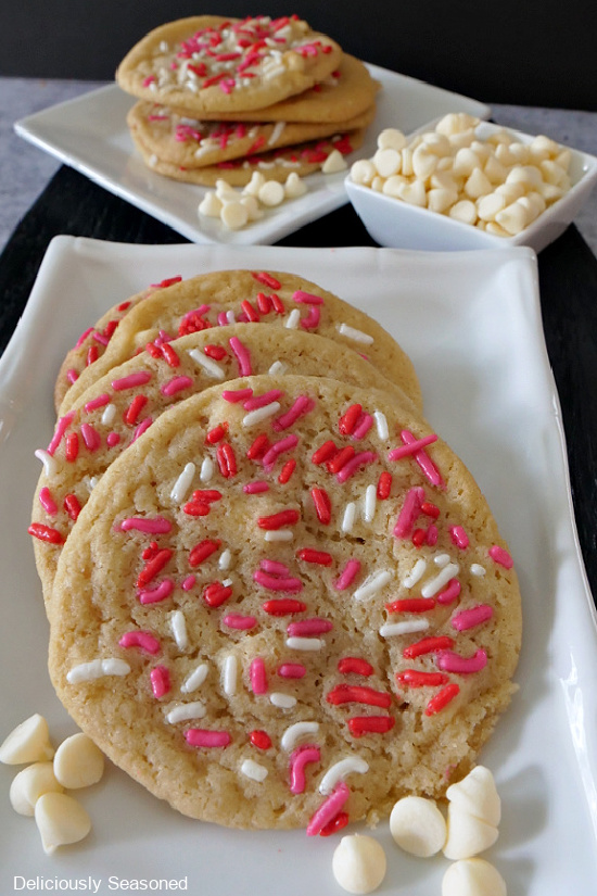 A white plate with 4 sugar cookies on it with another white plate in the background with 4 cookies stacked on it and a small white bowl of white chocolate chips.