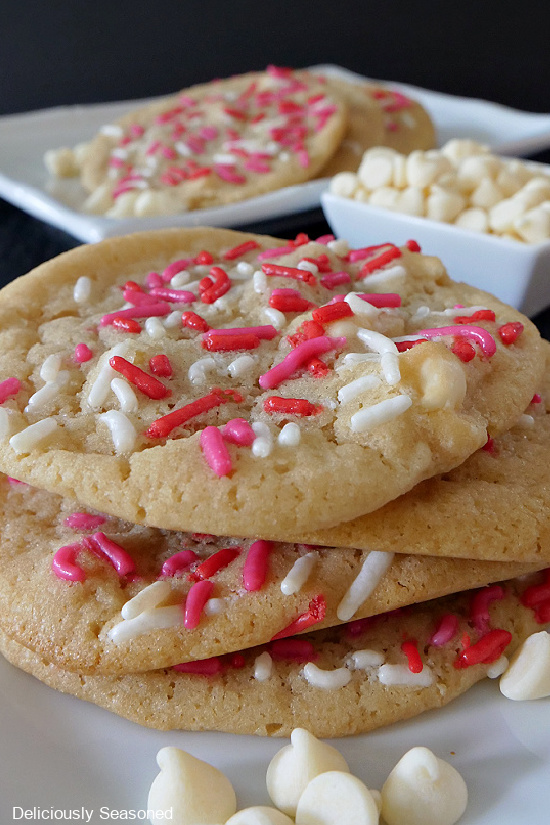 A white plate with 4 sugar cookies stacked with white chocolate chips scattered in the front, a small white bowl of white chocolate chips, and a white plate with another stack of cookies in the background.
