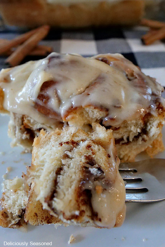 A bite of maple cinnamon roll on a fork that is laying on a white plate next to the cinnamon roll.