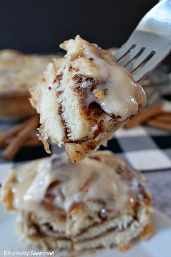 A bite of a maple cinnamon roll on a fork with the whole cinnamon roll underneath on a white plate.