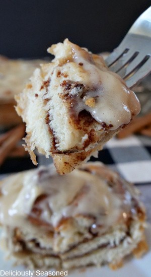 A close up picture of a bite of cinnamon roll on a fork with a white plate and a cinnamon roll in the background.