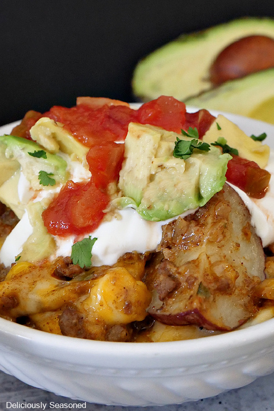 A close up of a white bowl filled with loaded roasted potatoes with sour cream, diced avocado and salsa on top.