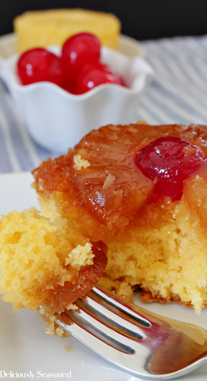 A close up photo of a slice of pineapple upside down cake with a bite sitting on a fork and a small bowl of cherries and pineapple slices in the background.