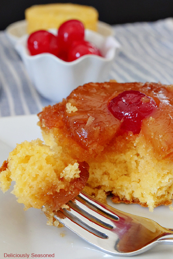 A close up of a piece of pineapple upside down cake on a white plate with a bite of cake on a fork laying on the plate.