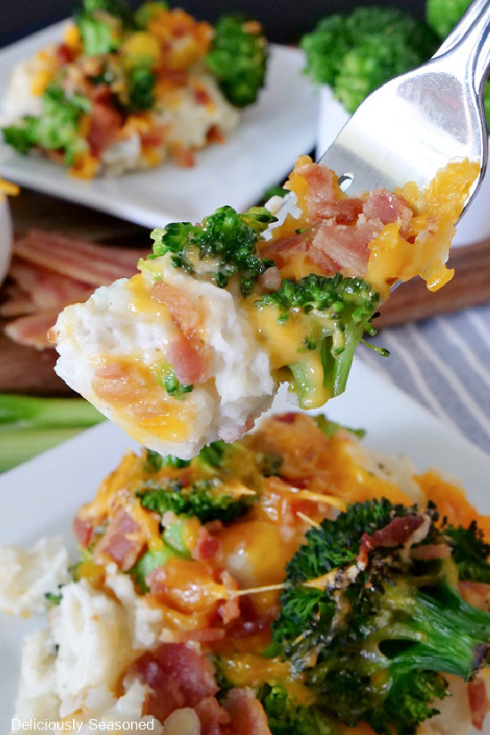 A fork loaded with a bite of broccoli mashed potato bake with a white plate filled with a serving of this side dish recipe and another white plate with a serving on it in the background.