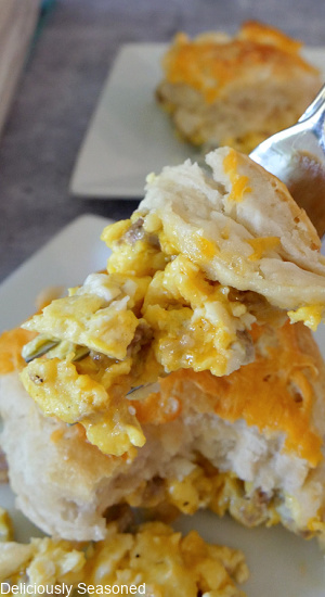 A white plate with a fork of scrambled eggs, sausage, cheese, topped with a biscuit.