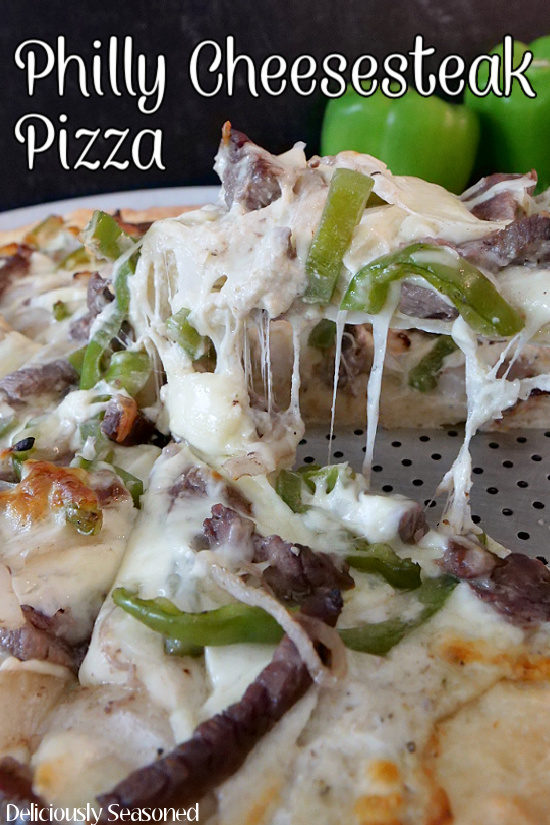 Philly cheesesteak pizza on a silver pizza pan, with green bell peppers in the background, and a slice being lifted off of the pan.