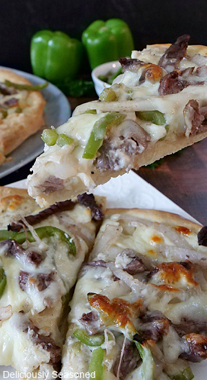 A white plate with Philly cheesesteak pizza and a piece being held up with a pizza pan and green bell peppers in the background.