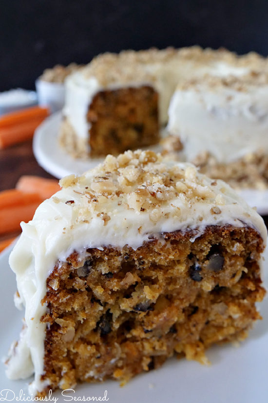 A close up shot of a slice of carrot cake with cream cheese frosting on a white plate with the cake in the background.