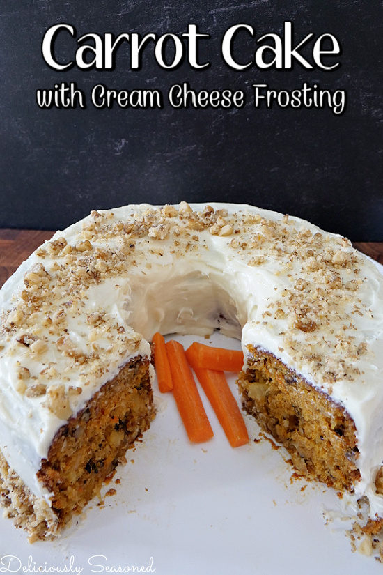 A carrot cake with cream cheese frosting on a white cake plate with a couple pieces removed with sliced carrots sitting on the plate in the middle of the cake.