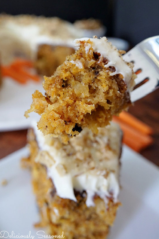 A bite of carrot cake with cream cheese frosting on a fork being held about a slice a cake that is on a white plate.