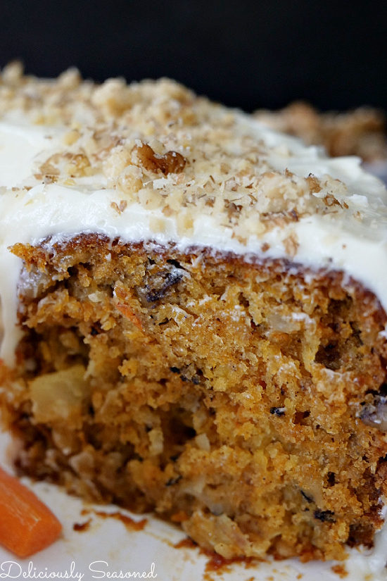 A close up view of carrot cake from the side view after a piece has been removed. It's on a white plate with cream cheese frosting and chopped walnuts of top.
