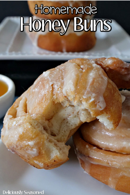 Two homemade honey buns stacked on a small white plate with a bite taken out of one and a bowl filled with honey and another white plate with honey buns on it in the background.