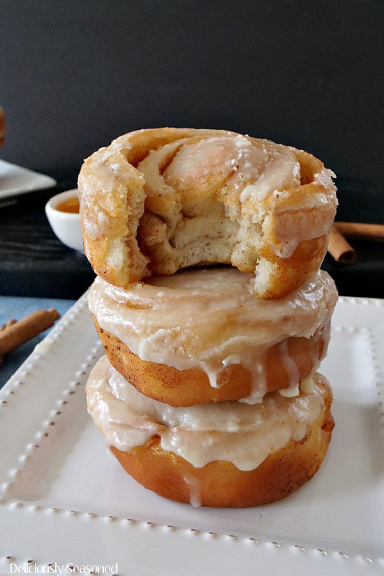 A stack of three honey buns stacked up on a white plate with a bite taken out of one, showcasing the delicious cinnamon center.