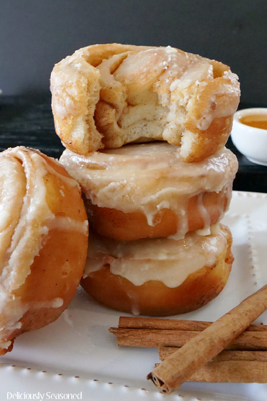 A close up image of 3 honey buns stacked on top of each other, with the top honey bun missing a bite, showcasing the delicious cinnamon center and another honey bun leaning up against the stack with cinnamon sticks laying on the white plate.