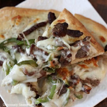 A slice of Philly cheesesteak pizza held above another slice of pizza on a white plate.