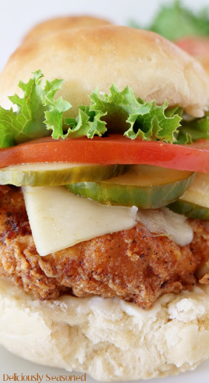 A close up picture of The Best Chicken Sandwich, showing the crispy fried chicken breast, melted cheese, homemade dill pickles, fresh tomato slice, and leafy green lettuce, all between a delicious, buttery homemade bun.