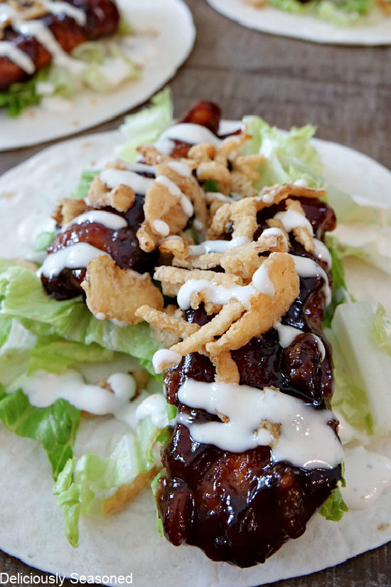 A close up photo of a flour tortilla with lettuce, honey BBQ chicken strips, crispy fried onion strings and ranch drizzled over the top.