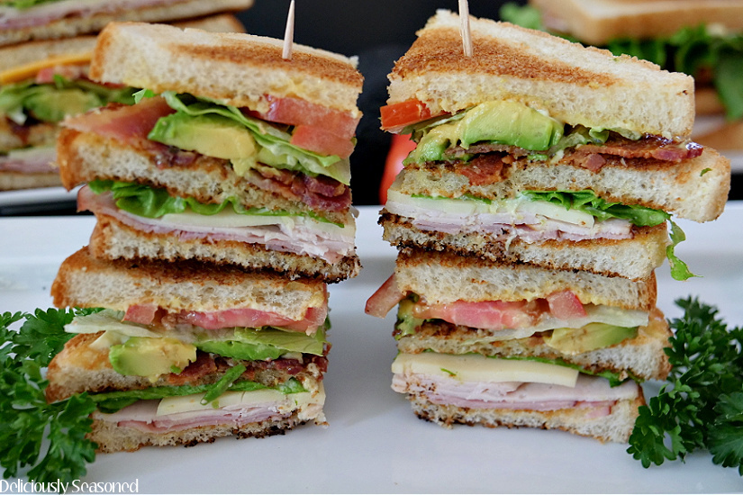 Club sandwiches cut into quarters and stacked two tall on a white plate with parsley for decoration. 