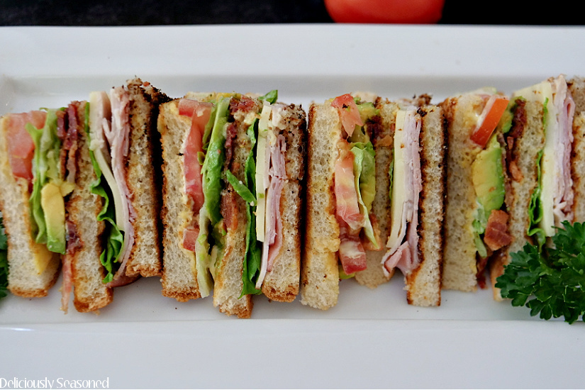 A horizontal photo of club sandwiches cut into quarters and lined up on a white plate, loaded with ham, turkey, avocado, bacon, lettuce, tomato, and cheese.