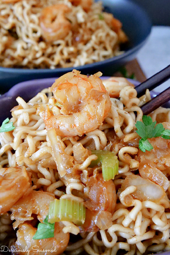 A purple bowl of shrimp chow mein with chopsticks lifting our a bite showing the ramen noodles and the shrimp.