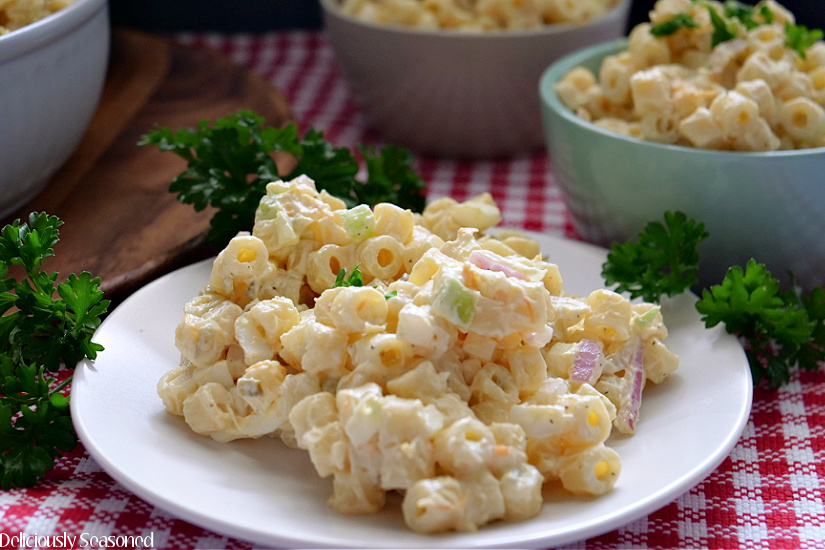 A white plate with a serving of macaroni salad on it with three bowls in the background filled with macaroni salad.