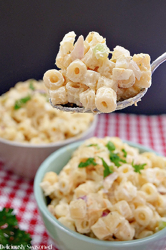 A close up photo of a spoonful of macaroni salad being held above a bowl filled with macaroni salad.