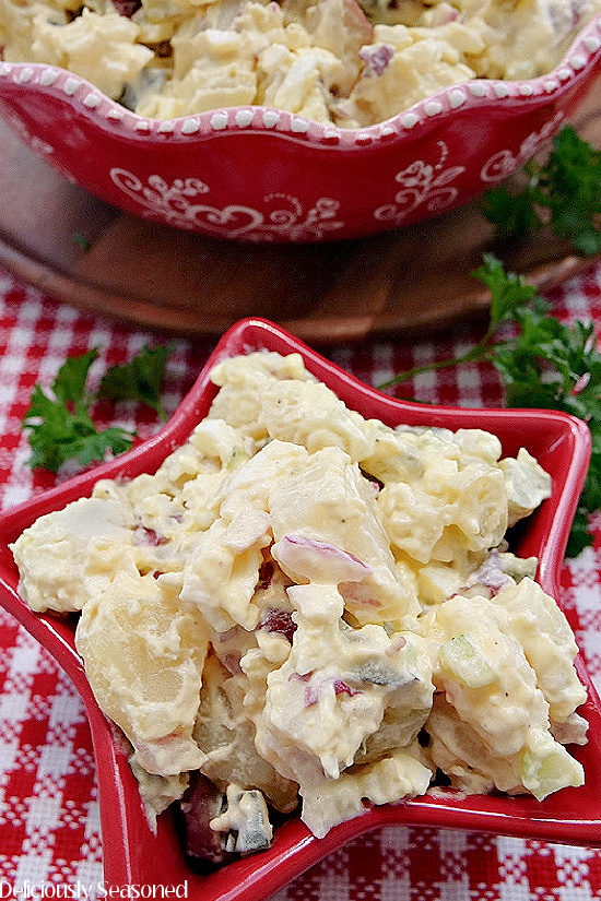 A close up photo of a red star bowl filled with potato salad that is sitting on a red and white checkered placemat.