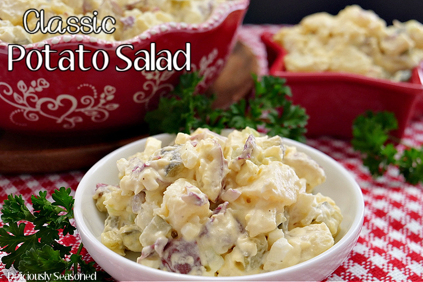 A white bowl filled with a serving of potato salad sitting on a red and white checkered placemat with a red star bowl and a big red bowl filled with potato salad in the background.