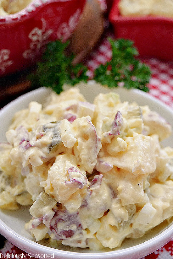 A close up photo of a white bowl filled with a serving of potato salad placed on a red and white checkered placemat with two red bowls in the background.