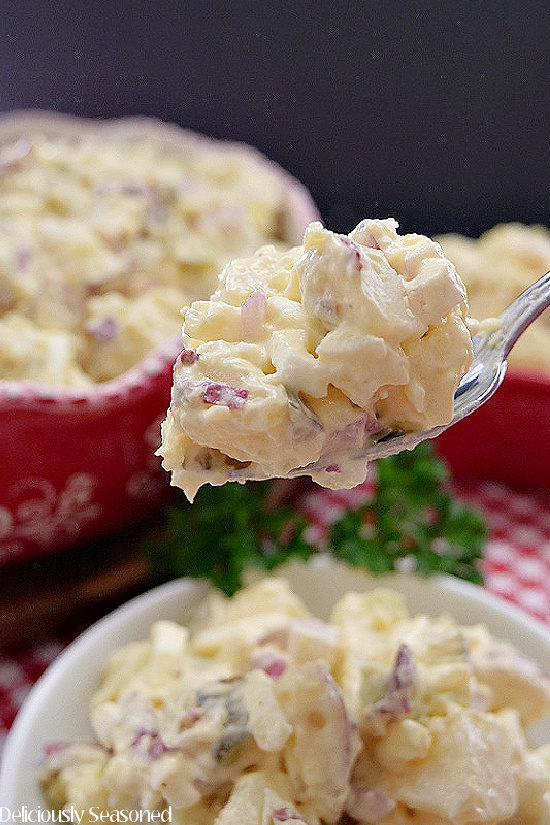 A close up picture of a big spoonful of potato salad being held above a white bowl filled with potato salad.