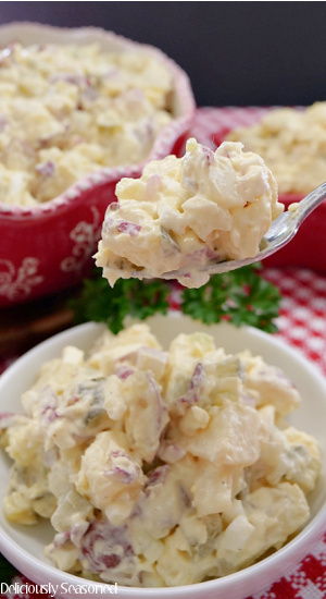 A close up photo of a spoonful of potato salad being held above a white serving bowl filled with potato salad also with a big red bowl filled with potato salad.