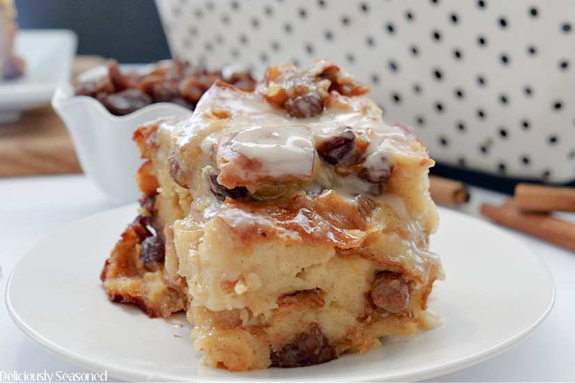 A piece of cinnamon raisin bread pudding on a white plate with a small white bowl of raisins and a large casserole dish in the background.